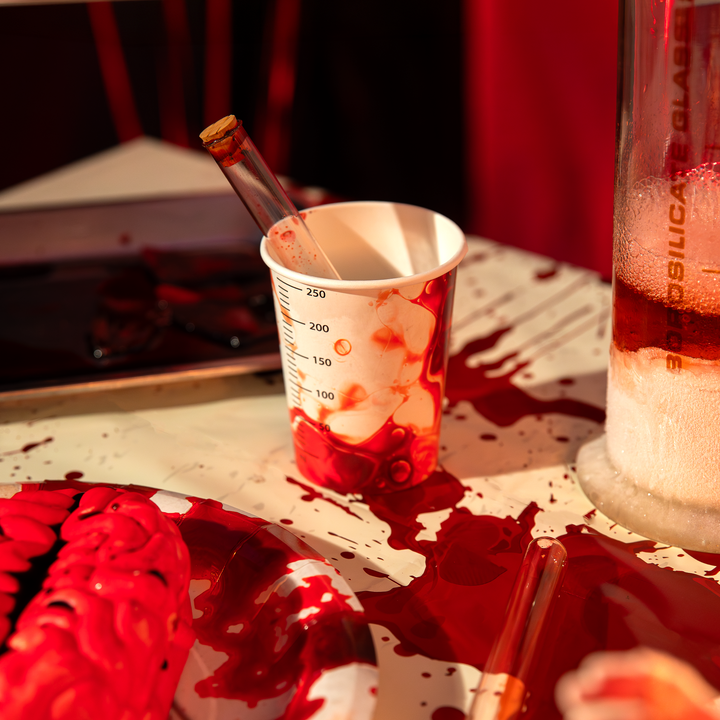 Kubek papierowy jednorazowy z motywem naczynia laboratoryjnego z miarką, z nadrukiem płynu przypminającym krew, z kolekcji bloody na halloween. w kubku znajduje się probówka laboratoryjna. Kubek stoi na stole przykrytym obrusem z tej samej kolekcji z motywem krwi