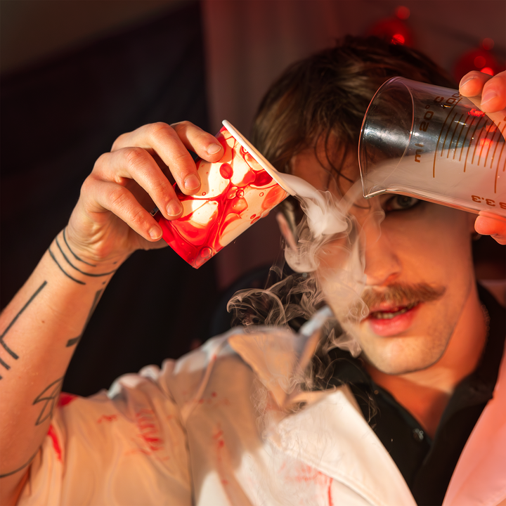 Chłopak trzyma w jednej ręce laboratoryjne naczynie z podziałką, a w drugiej jednorazowy papierowy kubek z kolekcji Bloody na Halloween, ozdobiony motywem naczynia laboratoryjnego z nadrukiem płynu przypominającego krew. Po przechyleniu obu naczyń wydobywa się gęsta mgła imitująca efekt suchego lodu