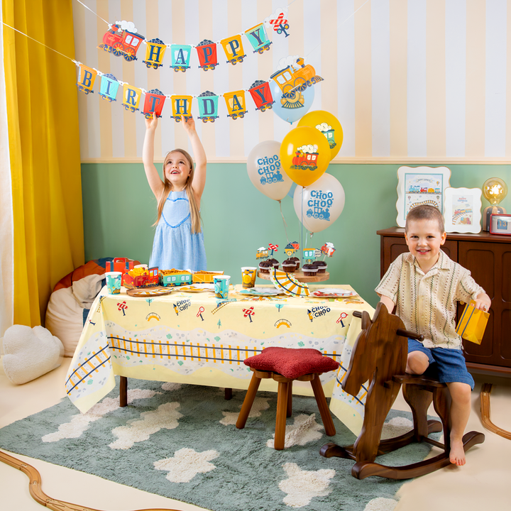 Dziecięca impreza z motywem ciuchci, uśmiechnięta dziewczynka trzymająca baner z napisem happy birthday, radosny chłopiec na koniku na biegunach trzymający torebkę na cukierki w kształcie  czerwonej walizki. Na stole talerzyki, kubeczki, serwetki, pudełka na chipsy, babeczki udekorowane topperami z motywem ciuchci, pociągów. za stołem balony lateksowe