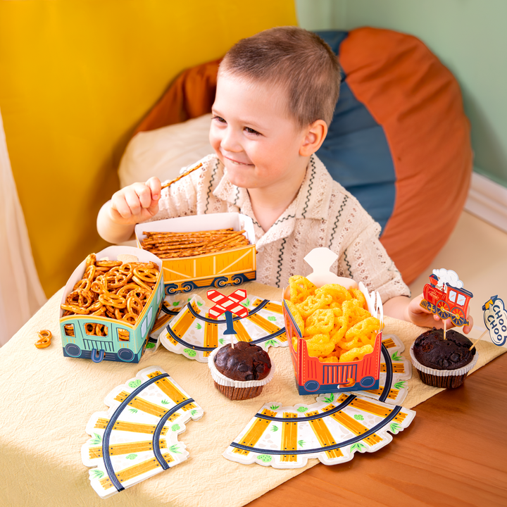 na stole pudełka na chipsy w kształcie lokomotywy z wagonikami z kolekcji ciuchcia, w pudełkach chipsy, paluszki oraz precelki, serwetki w kształcie torów kolejowych, babeczki udekorowane topperami z motywem pociągów. Chłopiec trzymający w ręku paluszki
