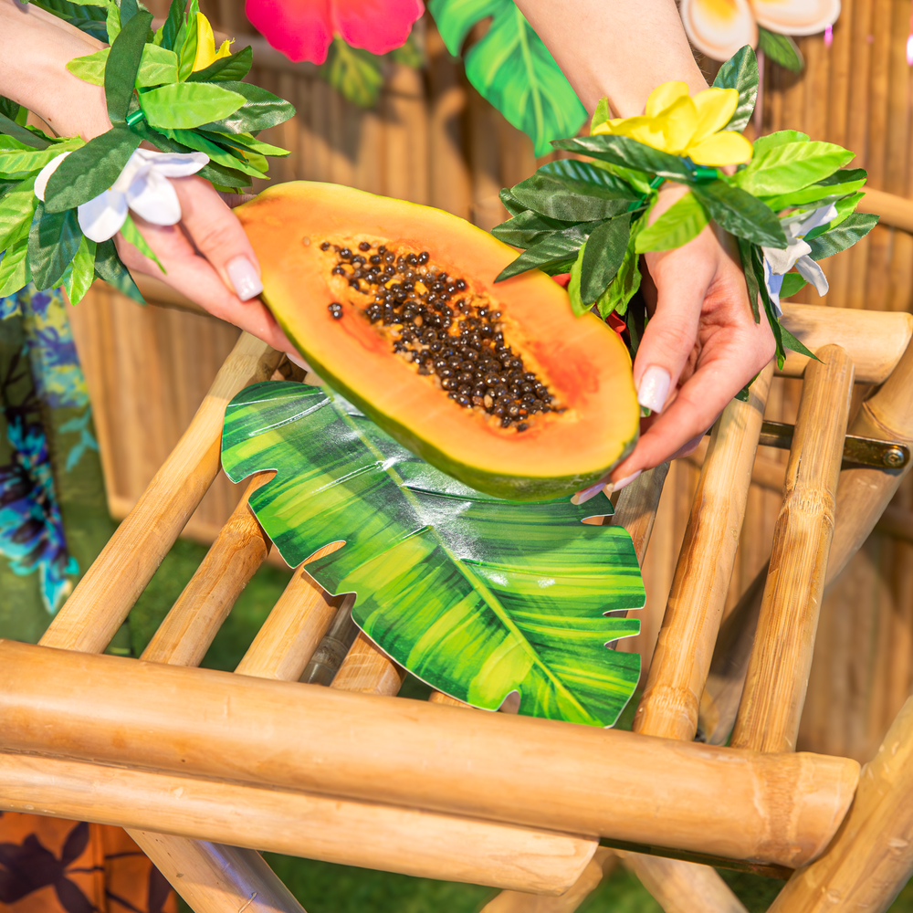 na bambusowym krześle papierowy jednorazowy talerz w kształcie liścia bananowca, papaja trzymana przez dziewczynę, na nadgarstkach bransoletki w kształcie tropikalnych kwiatów