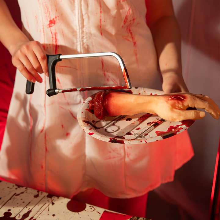 Dziewczyna przykłada piłę do sztucznej uciętej ręki, ktora leży na papierowym jednorazowym talerzu z motywem narzędzi chirurgicznych i krwi z kolekcji bloody na halloween