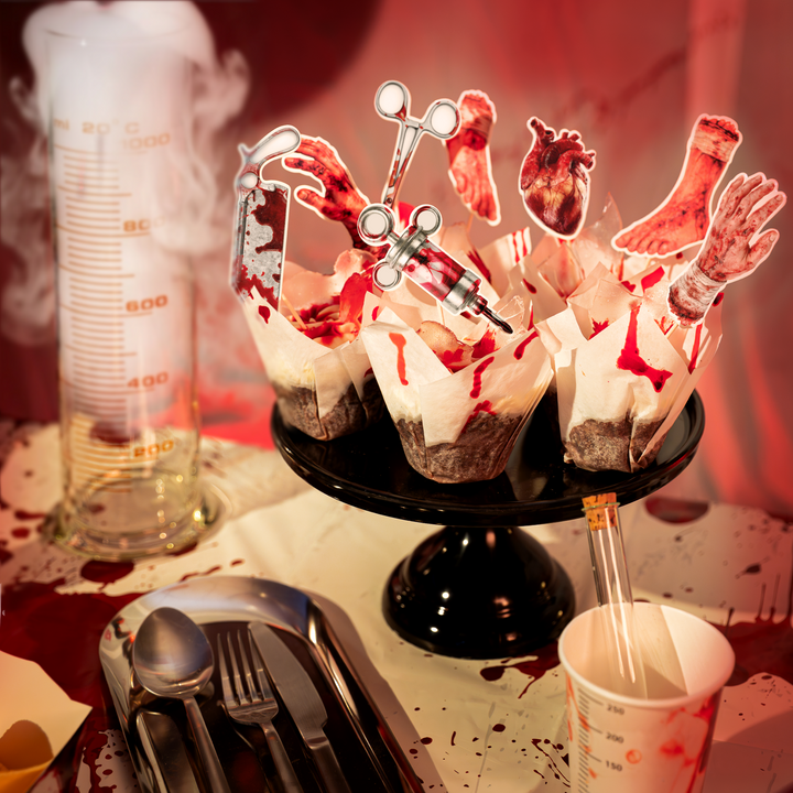 muffinki ozdbione topperami w kształcie strzykawki, tasaka, nożyczek chirurgicznych, zakrwawionych dłoni i stóp oraz serca z kolekcji bloody na halloween. Obok probówka w kubeczku papierowym oraz stalowe sztućce