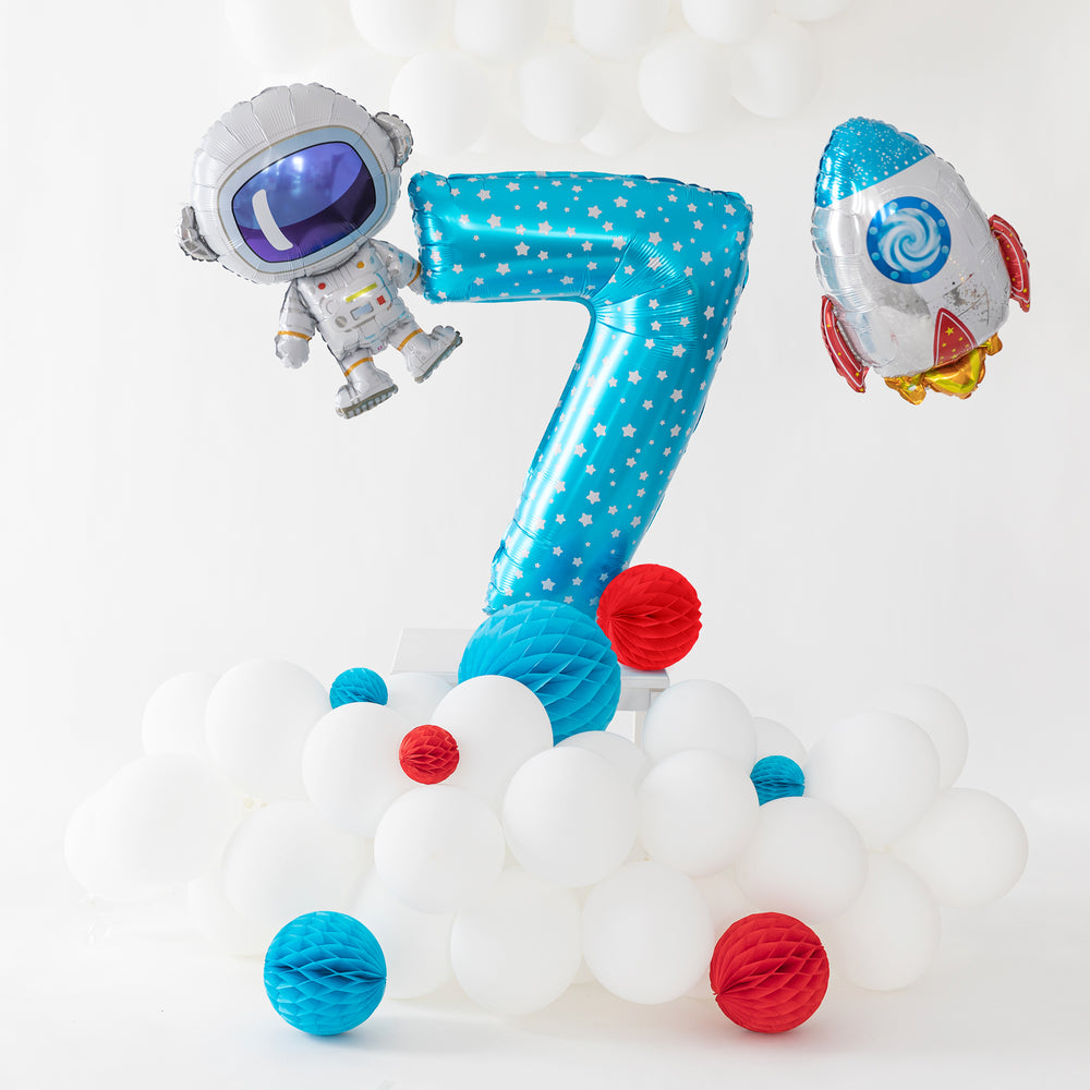 Na zdjęciu znajduje się balon cyfra 7 z balonami foliowymi w kształcie rakiety i kosmonauty oraz białymi balonami lateksowymi oraz białymi i niebieskimi honeycomb