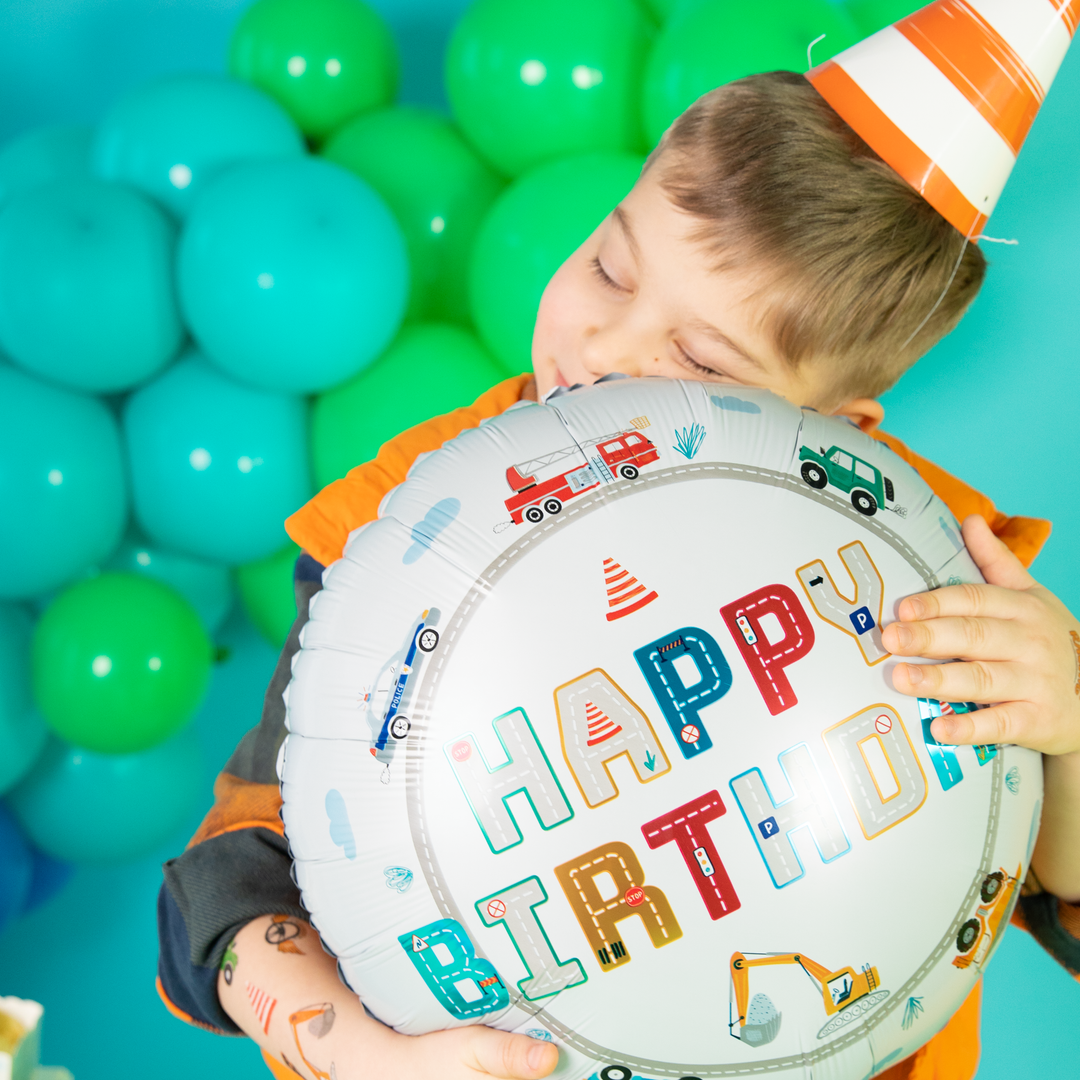 Chłopczyk przytula się do balonu foliowego z samochodzikami i napisem Happy Birthday.