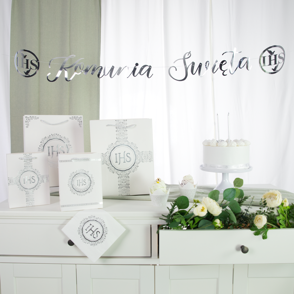 Srebrny baner komunijny na zdjęciu aranżacyjnym z torebkami na prezenty i serwetką