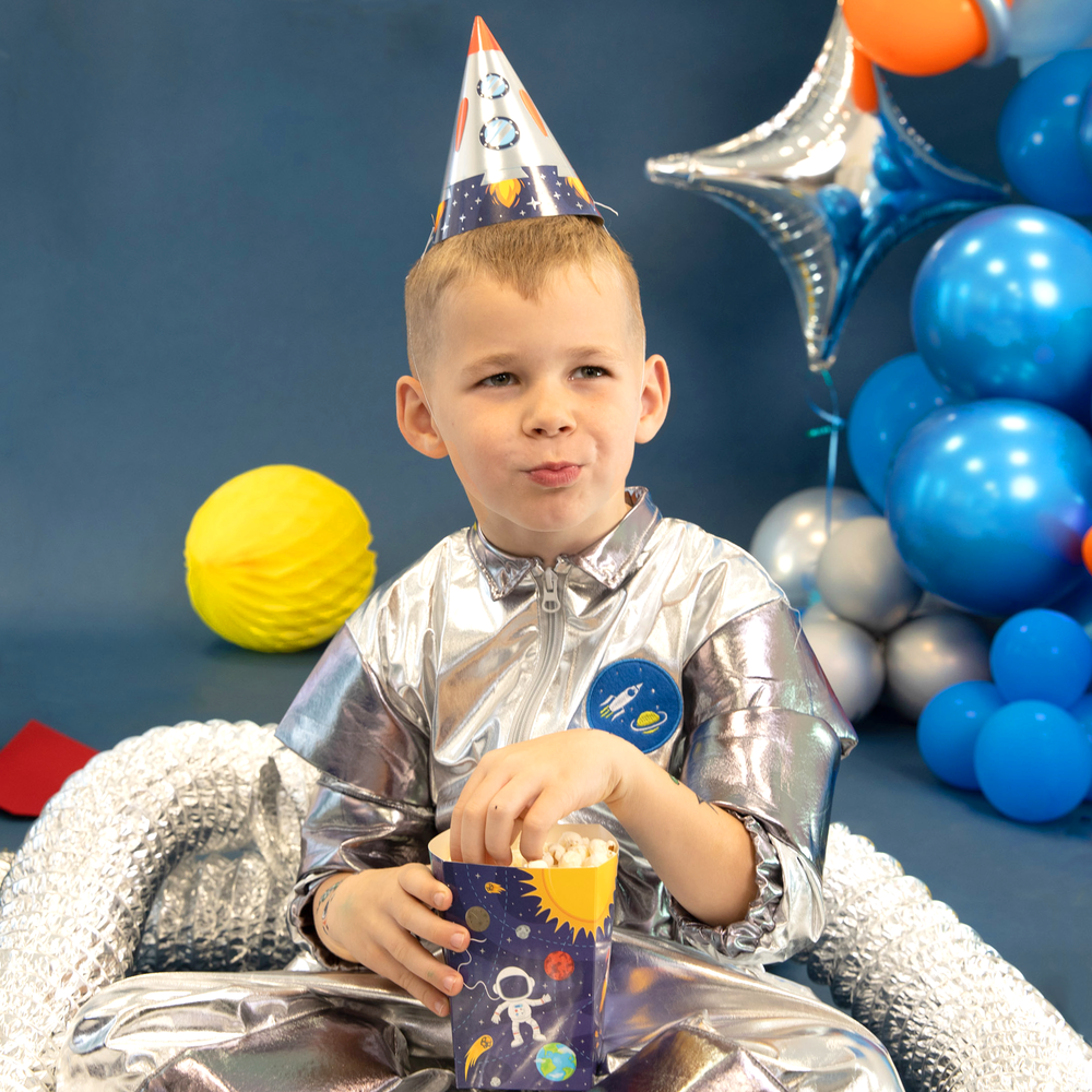 Chłopiec w srebrnym kombinezonie w czapeczce z rakietą na głowie, siedzi na podłodze i je popcorn z pudełka z kolekcji Kosmos