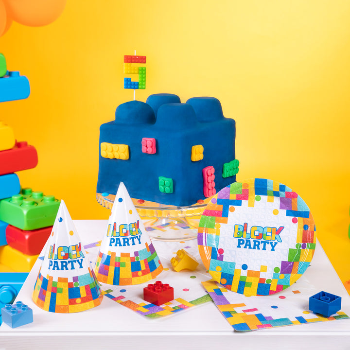Tort przypominający klocek ze świeczką cyfra 5, talerzykiem i czapeczkami imprezowymi z kolorową grafiką  przypominającą klocki lego.