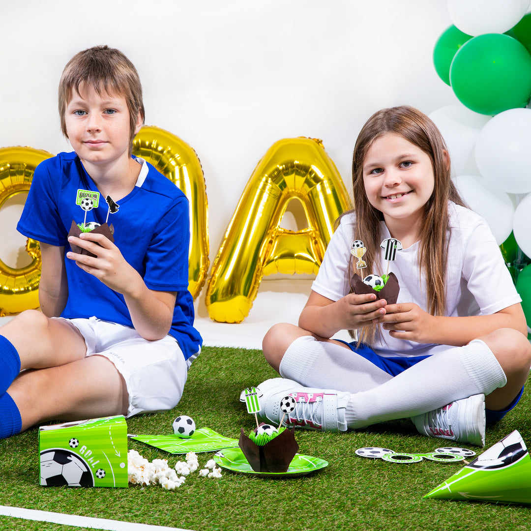 Dzieci siedzące na zielonym dywanie  trzymające babeczki z topperami a przed nimi znajdują się inne elementy z kolekcji Football.