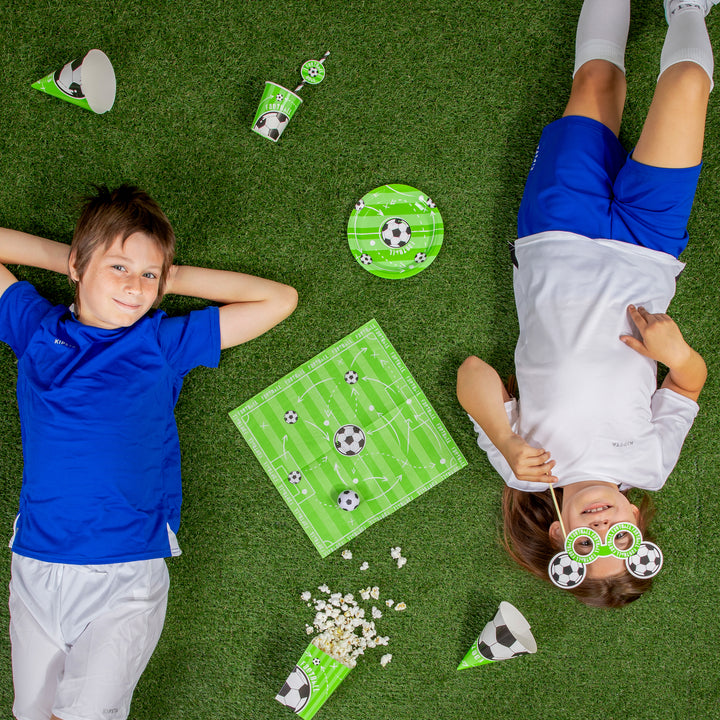 Dzieci leżące na zielonym dywanie z produktami z kolekcji Football  z centralnie ułożoną serwetka.