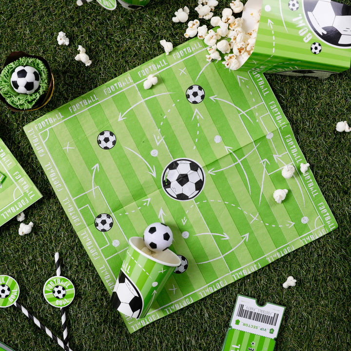 Zdjęcie serwetki, kubeczka, słomek i pudełka z kolekcji Football z rozsypanym na zielonej murawie popcornem.
