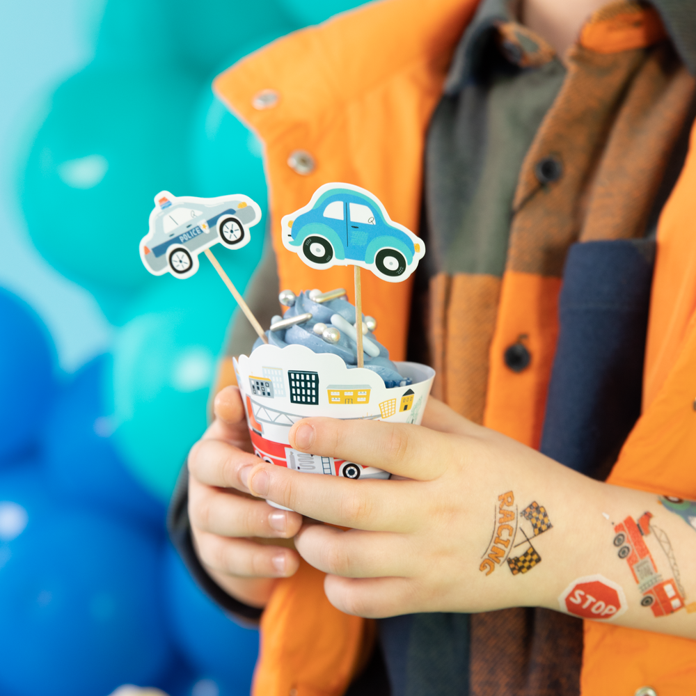 Chłopiec trzyma babeczkę w papilotce i z topperami z autami: garbusem i radiowozem