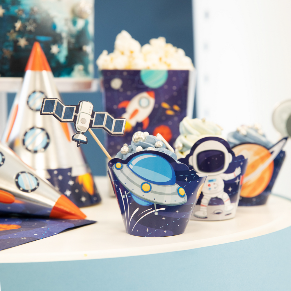 Zdjęcie babeczki w papilotce ze statkiem kosmicznym i topperem z satelitą na tle innych elementów z kolekcji Kosmos