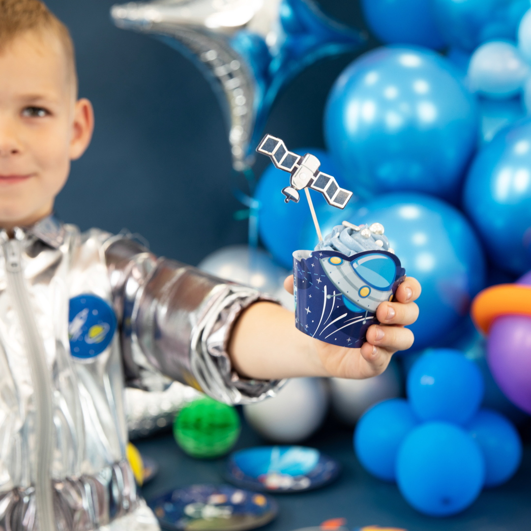 Chłopiec w stroju astronauty trzyma w rączce babeczkę w papilotce ze statkiem kosmicznym i topperem z satelitą