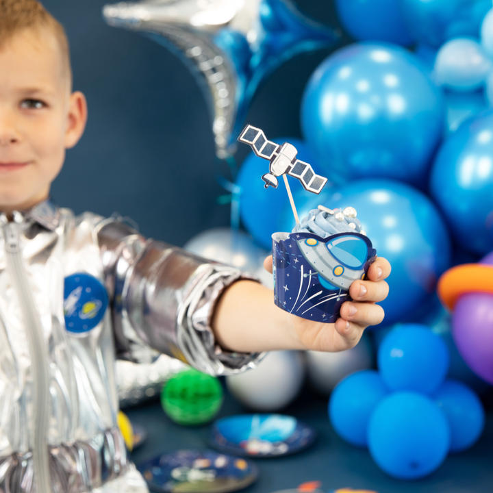 Chłopiec w stroju astronauty trzyma w rączce babeczkę w papilotce ze statkiem kosmicznym i topperem z satelitą